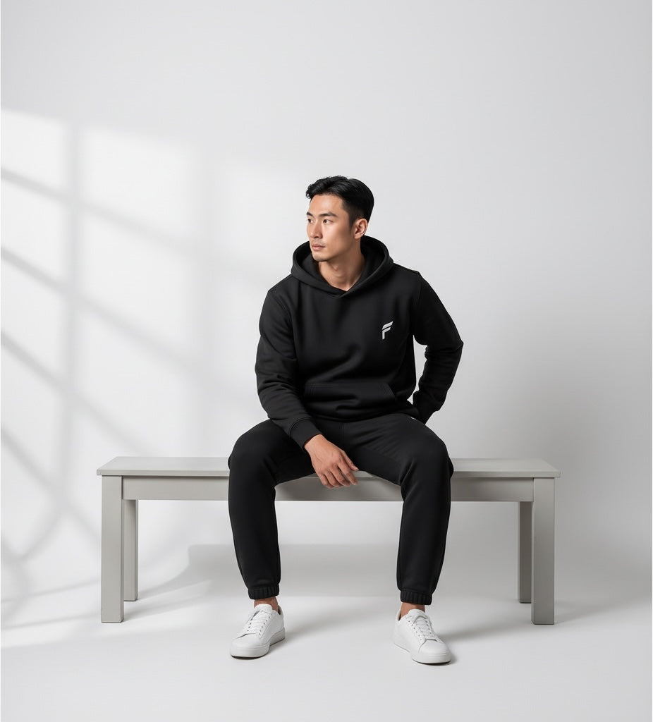 A man wearing a black FITTING fleece hoodie and matching joggers, sitting on a bench in a minimalist studio setting.