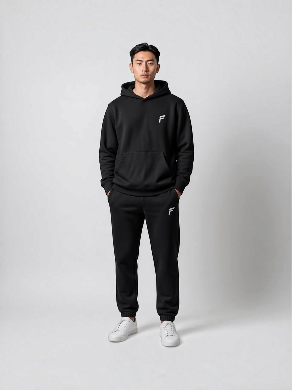 A man wearing a black FITTING Essential fleece hoodie and jogger set with white sneakers against a grey background.