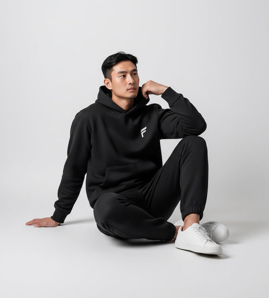 A man wearing a black FITTING fleece hoodie and matching joggers, featuring a minimalist logo and white sneakers.
