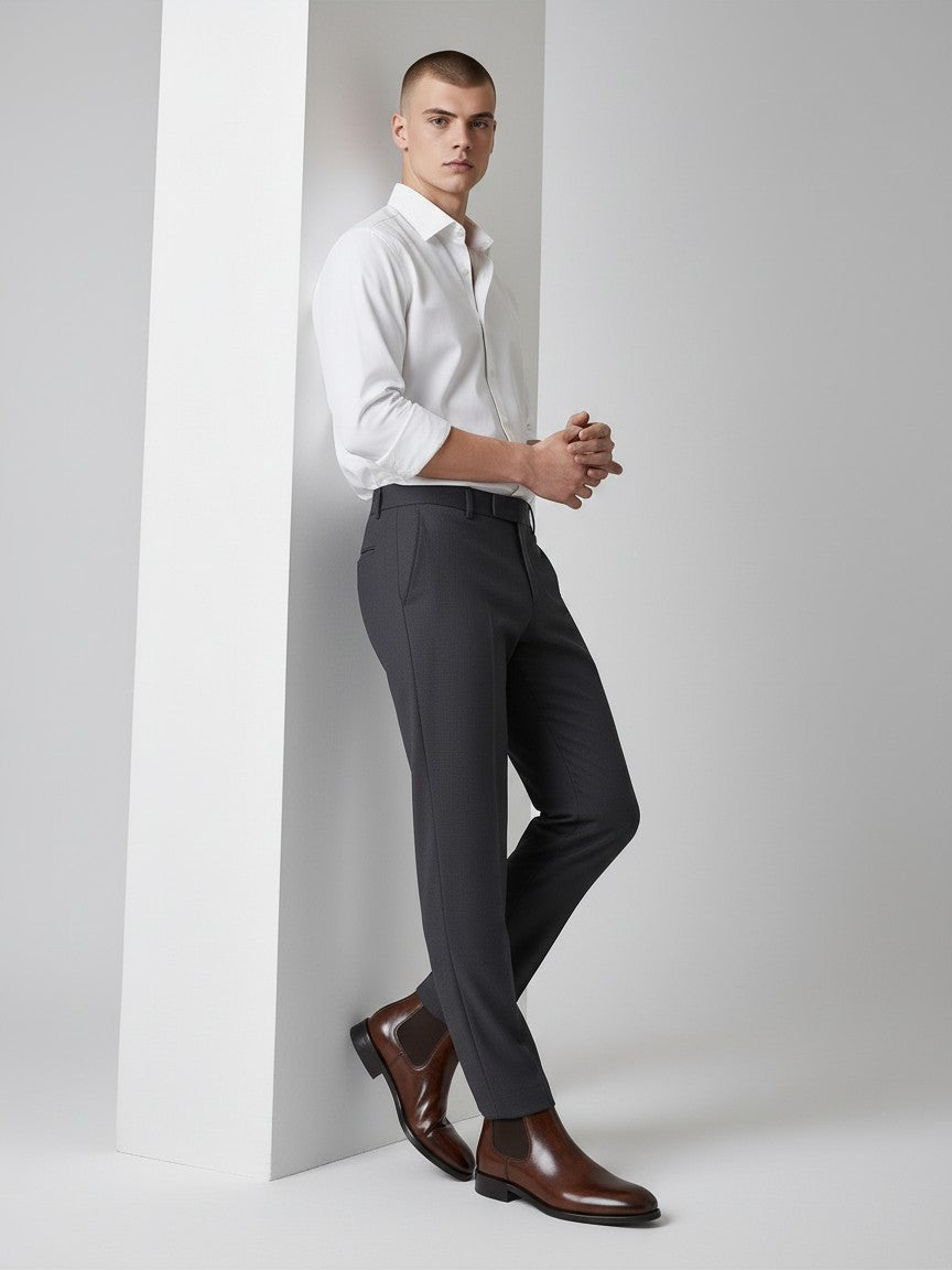 FITTING mahogany leather Chelsea boots worn by a man in a white shirt and charcoal trousers against a white pillar.