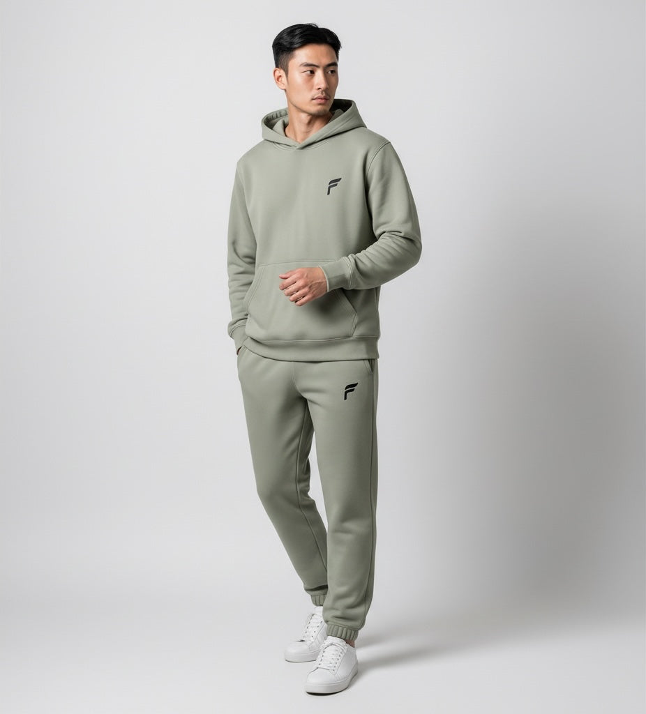A man wearing a sage green FITTING fleece hoodie and jogger set with a minimalist logo, paired with white sneakers.